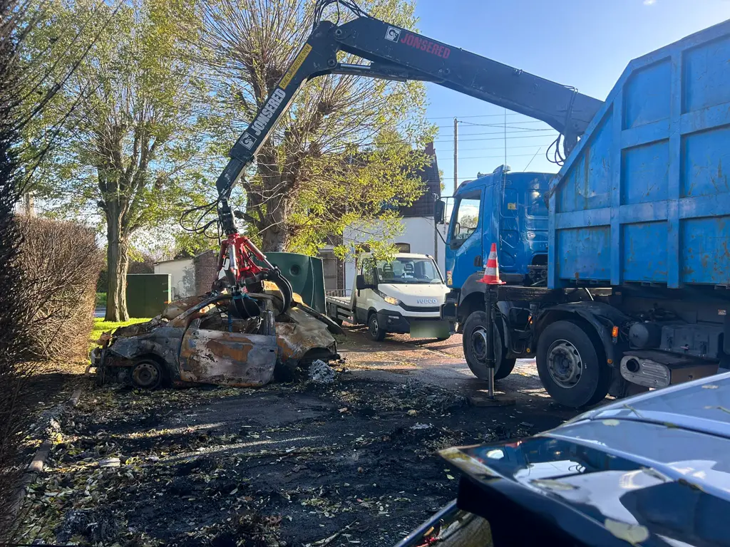 Un camion-grue soulève une voiture entièrement brûlée au bord d'une route ; une dépanneuse blanche attend derrière.